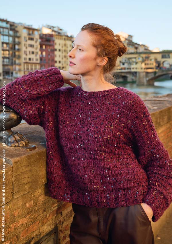 Femme accoudée sur un mur en pierre au bord d'une rivière, portant un pull en tricot bordeaux pailleté à grosses mailles torsadées et un pantalon en cuir marron, décor de Florence en arrière-plan.