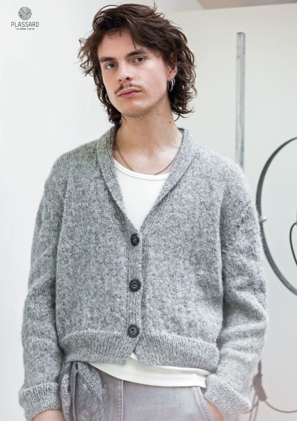 Homme aux cheveux bouclés portant un gilet cardigan en tricot gris chiné avec col châle et boutons foncés, sur un pantalon de jogging clair. Modèle Plassard.