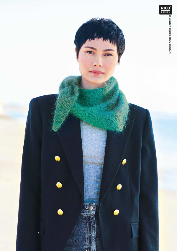 Femme aux cheveux courts portant un snood/écharpe tricoté en mohair vert, associé à un blazer marine à boutons dorés et un jean, sur fond de plage. Modèle Rico Design.