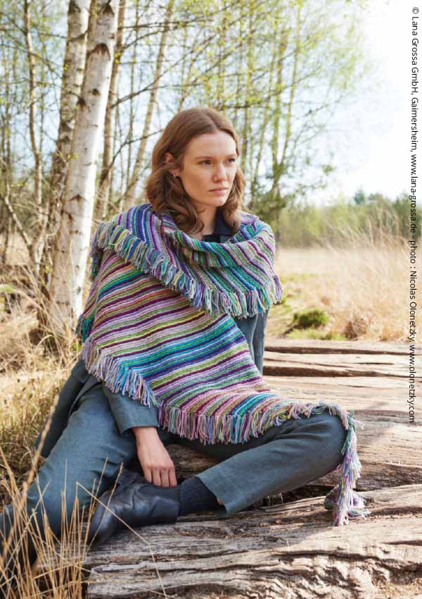Femme assise en tricot rayé multicolore façon poncho, dans un décor de bouleaux en extérieur.