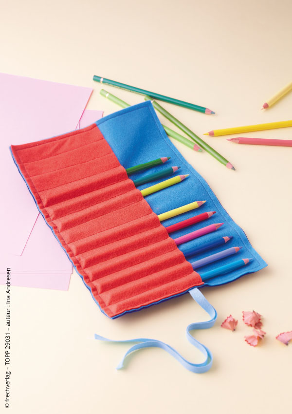 Trousse à crayons en tissu rouge et bleu, roulée et fermée par un ruban, entourée de crayons de couleur et de papier rose sur fond beige.