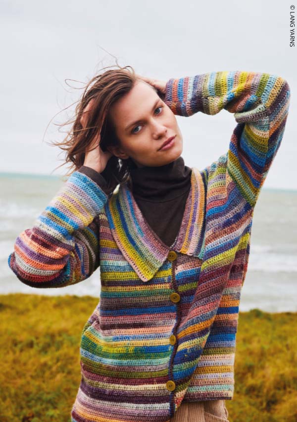 Femme portant un cardigan en crochet rayé multicolore, cheveux au vent, en bord de mer.