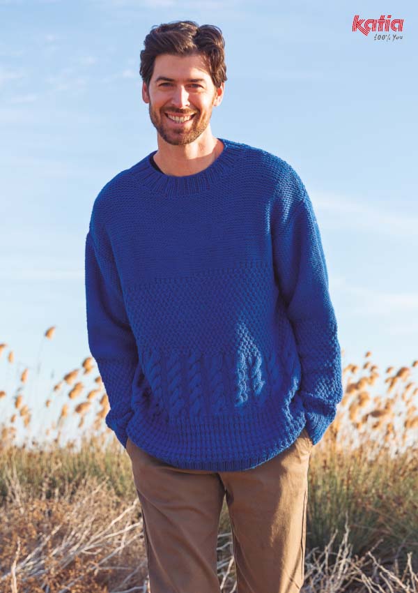 Homme portant un pull en tricot bleu roi à motif torsadé au niveau du poignet, dans un champ de roseaux.