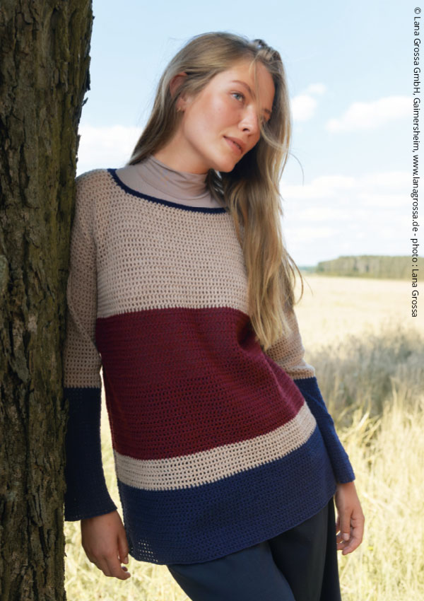 Femme portant un pull en crochet bicolore, beige et bordeaux, avec col bleu marine, devant un arbre en bord de plage.