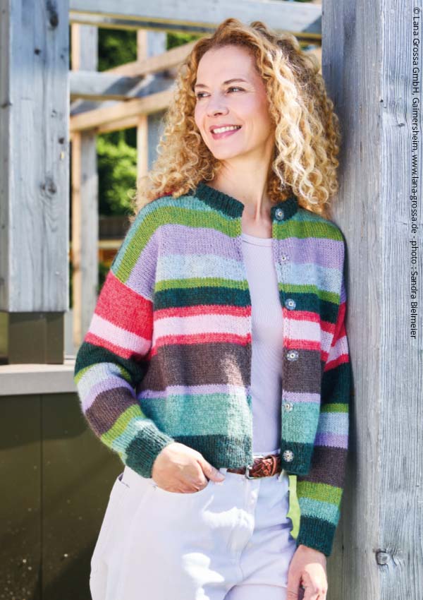 Femme aux cheveux bouclés portant un gilet en tricot rayé multicolore, boutonné, devant une façade en bois.