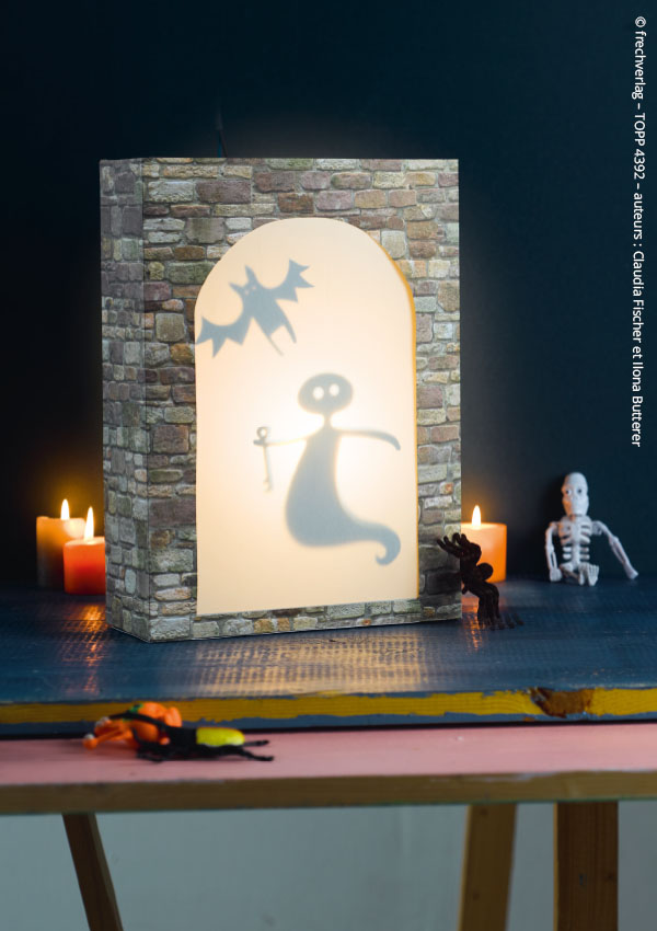 Lanterne en papier façon château hanté avec un fantôme lumineux, entourée de bougies allumées et de figurines squelette et insecte pour une décoration d'Halloween